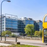 Yellow bus passes modern glass office buildings.