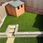 Fenced garden with grass, shed, and paved walkway.