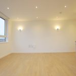 Modern empty room with wooden flooring and kitchen area.