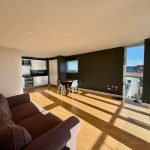 Modern open-plan living room with kitchen and large windows.