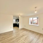 Modern empty room with wooden flooring and kitchen view.