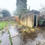 Overgrown backyard with stone shed and bins.