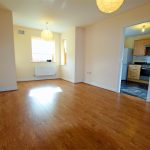 Empty room with wooden flooring and kitchen view.
